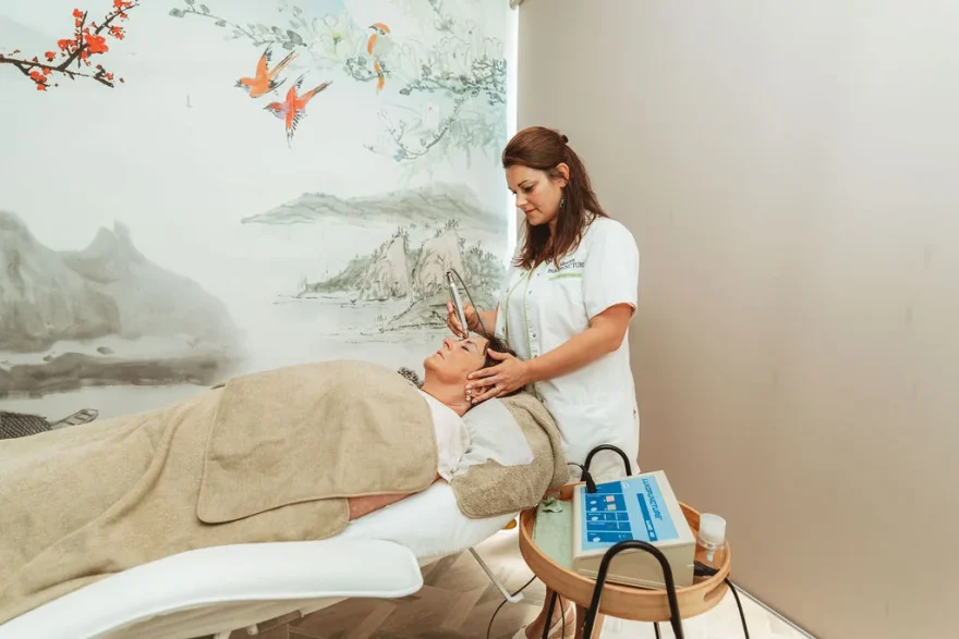 Praticienne en blouse posant un stylet de luxopuncture sur le front d’une cliente allongée, dans une cabine du cabinet de Rochefort