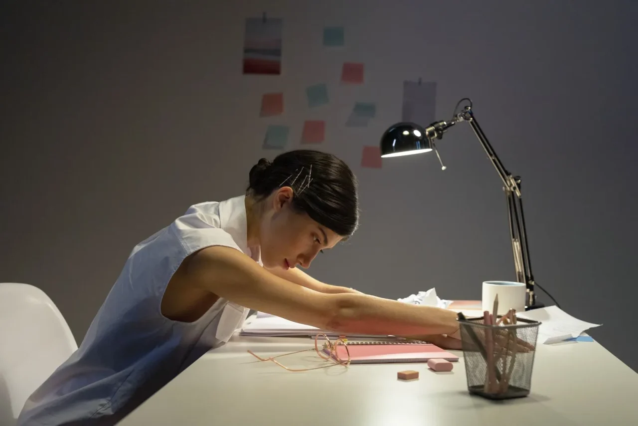 Une femme fatiguée se repose à son bureau, dans une pièce peu éclairée